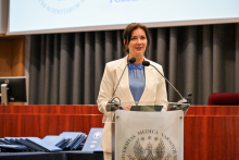 Kobieta w białej marynarce i niebieskiej bluzce stoi za mównicą z logo Warszawskiego Uniwersytetu Medycznego. Na mównicy są dwa mikrofony. 