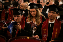 Grupa absolwentów siedzi w audytorium. Mają czarne togi i birety z emblematami uczelni. Na czerwonych szarfach widnieje napis „CLASS OF 2025” i „GRADUATE”. Niektórzy trzymają programy lub broszury. W tle widać innych uczestników ceremonii.