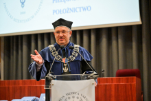 Mężczyzna w niebiesko-czarnej todze, z czarnym biretem na głowie, w okularach, prof. Gałązkowski, stoi za mównicą i przemawia. Prawą rękę ma uniesioną na wysokość piersi, uśmiecha się. Za nim ekran projekcyjny.