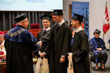 Trzech absolwentów ratownictwa medycznego w czarnych togach i biretach odbierają gratulacje od wykładowcy w czarno-niebieskiej todze z łańcuchem akademickim. Trzymają torby z logo uczelni. Uśmiechają się. W tle uczestnicy ceremonii, czerwono-biało-złoty sztandar uczelni i rollup.