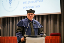 Mężczyzna w okularach, prodziekan Rzońca, w niebiesko-czarnej todze i birecie stoi za mównicą. Przemawia, lekko się uśmiecha.