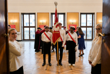 Troje studentów w białych koszulach i czarnych spodniach stoi na środku sali z drewnianą podłogą. Jeden trzyma czerwoną flagę z białym orłem. W tle osoby w togach i dwie osoby przy drzwiach. Sala ma jasne ściany, duże okna i lampy na ścianach.