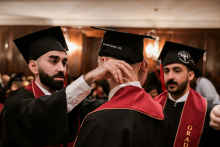 Trzech absolwentów w togach i biretach podczas ceremonii. Dwóch ma czerwone szarfy z napisem „GRADUATION”. Jeden poprawia biret drugiemu. W tle drewniane panele ścienne i lampy.