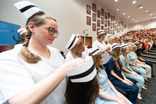Uroczystość czepkowania absolwentów pielęgniarstwa i położnictwa. Starsze kobiety w białych uniformach zakładają czepki pielęgniarskie na głowy młodych kobiet siedzących w rzędzie. 