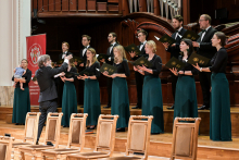 Chór mieszany występuje w sali koncertowej. Dyrygent stoi przodem do chóru z uniesionymi rękami. Kobiety mają zielone spódnice i czarne bluzki, mężczyźni czarne garnitury z muszkami. W tle duży instrument i czerwony baner z logo „Warszawski Uniwersytet Medyczny”. Na pierwszym planie puste drewniane krzesła.
