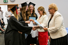 Trzy młode kobiety w czarnych togach i biretach odbierają dyplomy od dwóch kobiet. Jedna kobieta ma biały żakiet i czerwoną spódnicę, druga biały żakiet i czarne spodnie. W tle uczestnicy i zegar wskazujący 10:10.