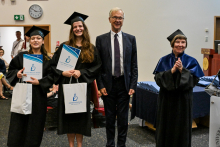 Cztery osoby pozują do zdjęcia. Dwie absolwentki w czarnych togach i biretach trzyma dyplomy i białe torby. Pośrodku stoi mężczyzna w garniturze. Po lewej stronie mężczyzna stoi kobieta w niebiesko-czarnej todze akademickiej i klaszcze. W tle widoczne są inne osoby, zegar na ścianie i stół z dyplomami.