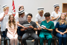 Uroczystość czepkowania absolwentów pielęgniarstwa i położnictwa. W pierwszym rzędzie siedzą cztery osoby w kolorowych uniformach medycznych (dwie młode kobiety i dwóch młodych mężczyzn). Za nimi stoją cztery osoby w białych uniformach, które zakładają siedzącym czepki pielęgniarskie na głowy siedzących.