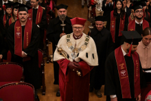 Uroczystość akademicka. W centrum osoba w czerwonym stroju i białym płaszczu z insygniami rektorskimi, trzyma berło. Po bokach absolwenci w czarnych togach i biretach, z bordowymi szarfami „CLASS OF 2025 GRADUATE”. W tle uczestnicy ceremonii.