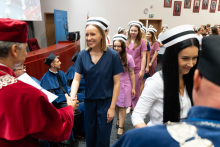 Uroczystość czepkowania absolwentów pielęgniarstwa i położnictwa. Młode kobiety w uniformach medycznych i czepekach pielęgniarskich stoją w kolejce do starszego mężczyzny w czerwonej todze, który wręcza im dyplomy. 