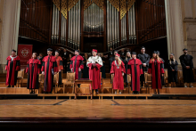 Grupa osób w czerwonych i czarnych togach oraz biretach stoi na scenie w sali koncertowej. Na środku osoba w białej todze z łańcuchem rektorskim. W tle duże srebrne organy piszczałkowe. Podłoga drewniana, widoczne ozdobne elementy architektoniczne.