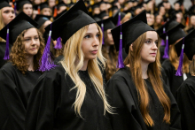 Grupa absolwentów w czarnych togach i biretach z fioletowymi chwostami stoi w rzędach. W tle widoczne są kolejne osoby ubrane podobnie. Wszyscy mają poważne twarze, powtarzają słowa ślubowania.