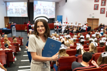 Uroczystość czepkowania absolwentów pielęgniarstwa i położnictwa. Młoda kobieta w beżowym uniformie medycznym i czepku pielęgniarskim stoi na środku audytorium, trzymając niebieską teczkę z napisem „Warszawski Uniwersytet Medyczny”. 