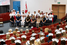 Uroczystość czepkowania absolwentów pielęgniarstwa i położnictwa. Grupa młodych kobiet w różnokolorowych uniformach medycznych i czepkach pielęgniarskich siedzi na scenie w audytorium. Za nimi stoją starsze kobiety w białych uniformach, zakładające czepki pielęgniarskie na głowy siedzących.