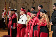 Grupa osób w togach akademickich stoi na scenie. Na pierwszym planie osoba w czerwonej todze z białym futrem i berłem. Pozostali mają czerwone togi z czarnymi elementami i czarne birety. W tle mężczyźni w białych koszulach z flagami. Scena na tle marmurowej ściany.