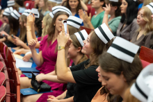 Uroczystość czepkowania absolwentów pielęgniarstwa i położnictwa. Grupa młodych kobiet w różnokolorowych uniformach medycznych i czepkach pielęgniarskich siedzi w audytorium. 