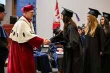 Mężczyzna w w czerwonym stroju akademickim z białym kołnierzem podaje rękę absolwentce w czarnej todze i birecie z fioletowym chwostem. W tle czerwono-biało-złoty sztandar uczelni i osoby w togach siedzące na krzesłach.