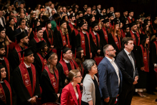 Grupa absolwentów w czarnych togach i biretach stoi podczas ceremonii. Mają czerwone stuły z napisem „CLASS OF 2025”. W tle widoczni są siedzący goście.