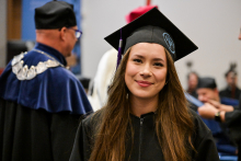 Młoda kobieta w czarnej todze i birecie stoi na pierwszym planie. Ma długie, brązowe włosy. W tle widoczne są inne osoby w togach oraz elementy sali akademickiej.