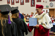 Dwie osoby w czarnych togach i biretach z fioletowymi chwostami stoją naprzeciw mężczyzny w czerwonej todze i birecie, który trzyma niebieski dyplom. W tle siedzą uczestnicy. Na ścianie widoczne są portrety w czerwonych ramkach i zegar.