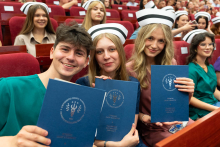 Uroczystość czepkowania absolwentów pielęgniarstwa i położnictwa. Młode kobiety i młody mężczyzna w uniformach medycznych i czepkach pielęgniarskich siedzą w audytorium, trzymając niebieskie teczki z napisem „Warszawski Uniwersytet Medyczny.”