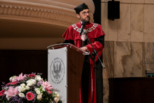 Mężczyzna w todze i birecie stoi przy mównicy z herbem i napisem „Uniwersytet Medyczny im. Karola Marcinkowskiego w Poznaniu”. Ma na szyi łańcuch rektorski. Przed mównicą bukiet z różowych, białych i fioletowych kwiatów. W tle marmurowa ściana i ozdobny gzyms.