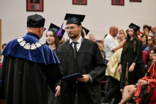 Ceremonia wręczenia dyplomów. Absolwent w czarnej todze i birecie odbiera dyplom od osoby w todze z niebieskim kołnierzem i łańcuchem rektorskim. W tle siedzą inni absolwenci i goście.