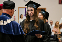 Ceremonia wręczenia dyplomów. Studentka w czarnej todze i birecie trzyma niebieską teczkę z dyplomem. Przed nią stoi osoba w granatowej todze z ozdobnym łańcuchem. W tle widoczni są absolwenci i goście.