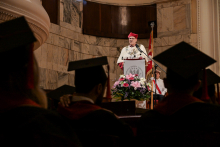 Uroczystość akademicka. W centrum mówca w białej szacie i czerwonym birecie za mównicą z herbem uczelni. Przed nim bukiet kwiatów. W tle marmurowy mur i flaga. Na pierwszym planie sylwetki absolwentów w czarnych togach i biretach, siedzących tyłem.