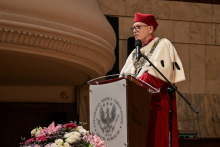 Mężczyzna w czerwonym birecie i białym płaszczu z czarnymi elementami stoi za mównicą z logo Uniwersytetu Medycznego w Warszawie. Przed mównicą bukiet różowych, białych i fioletowych kwiatów. W tle marmurowa ściana i ozdobny gzyms.