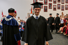 Ceremonia rozdania dyplomów. Absolwent w czarnej todze i birecie idzie w stronę aparatu. Po lewej stronie stoi rektor w granatowej todze z insygniami. Po prawej widoczni są inni absolwenci i publiczność. Na ścianie portrety w czerwonych ramkach.