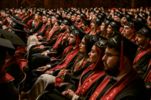 Duża grupa absolwentów w czarnych togach i biretach siedzi w audytorium. Mają czerwone szarfy z napisem „Class of 2025”. Sala jest pełna. W tle widoczna publiczność na balkonach.