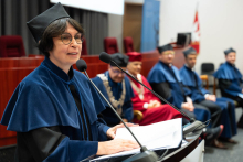 Uroczystość czepkowania absolwentów pielęgniarstwa i położnictwa. Kobieta w niebieskiej todze i birecie przemawia przy mównicy z dwoma mikrofonami. W tle siedzą cztery osoby w togach.