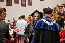 Absolwentka w czarnej todze i birecie odbiera dyplom od dziekana w niebiesko-czarnej todze z ozdobnym łańcuchem. W tle widać uczestników ceremonii i publiczność na trybunach. Ściana za nimi ozdobiona jest portretami.