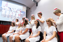 Uroczystość czepkowania absolwentów pielęgniarstwa i położnictwa. Starsze kobiety w białych uniformach i czepkach położnych stoją za rzędem młodych kobiet siedzących na krzesłach, również ubranych w białe uniformy i czepki położnych.