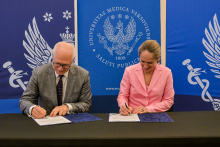 Mężczyzna w szarym garniturze i kobieta w różowym żakiecie podpisują dokumenty przy stole.