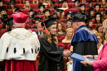 Ceremonia wręczenia dyplomów. Absolwentka w czarnej todze i birecie odbiera dyplom od osoby w czerwonym stroju akademickim. W tle inni absolwenci w togach i biretach siedzą w czerwonych fotelach.