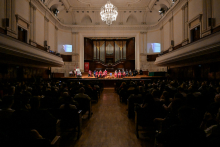 Duża sala koncertowa z wysokim sufitem i żyrandolem. Na scenie osoby w czerwonych togach siedzą na krzesłach. W tle duży organ piszczałkowy. Po bokach sceny ekrany pokazujące osobę przemawiającą przy mównicy. Publiczność siedzi w ciemności.