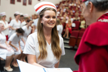 Uroczystość czepkowania absolwentów pielęgniarstwa i położnictwa. Kobieta w białym uniformie medycznym i czepku położnych odbiera dokument od starszego mężczyzny w czerwonym stroju akademickim.