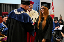 Kobieta w czarnej todze i birecie z rudymi włosami odbiera dyplom od mężczyzny w todze z niebieskim kołnierzem i łańcuchem. W tle uczestnicy ceremonii oraz czerwono-biało-złoty sztandar uczelni i rollup w kolorze niebieskim.