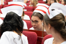 Uroczystość czepkowania absolwentów pielęgniarstwa i położnictwa. Grupa młodych kobiet w białych uniformach medycznych i czepkach położnych siedzi na czerwonych krzesłach w audytorium. Kobiety są zwrócone twarzami do siebie i rozmawiają.