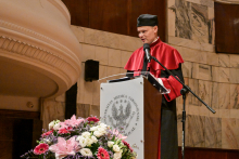 Mężczyzna w todze i birecie stoi za mównicą z logo Uniwersytetu Medycznego w Warszawie. Przed nim mikrofon i bukiet kwiatów w odcieniach różu, bieli i czerwieni. W tle marmurowe ściany i drewniane panele.