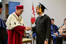 Młody mężczyzna w czarnej todze i birecie z jasnymi włosami odbiera dyplom od mężczyzny w todze w kolorach białym i czerwonym. W tle uczestnicy ceremonii oraz czerwono-biało-złoty sztandar uczelni i rollup w kolorze niebieskim.