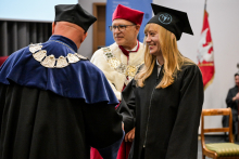 Absolwentka z jasnymi długimi włosami, w czarnej todze i birecie podaje rękę dziekanowi w niebiesko-czarnym stroju akademickim z łańcuchem. Za nimi rektor w czerwonej todze z białym kołnierzem i łańcuchem. Po prawej czerwono-biało-złoty sztandar uczelni.