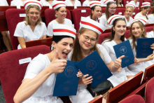Uroczystość czepkowania absolwentów pielęgniarstwa i położnictwa. Trzy kobiety w białych uniformach medycznych i czepkach położnych siedzą w rzędach czerwonych krzeseł. Dwie z nich pokazują kciuk w górę, a wszystkie trzy trzymają w rękach granatowe teczki. 