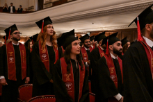 Grupa studentów w czarnych togach i biretach stoi w rzędach podczas ceremonii ukończenia studiów. Na czerwonych szarfach widnieje napis „CLASS OF 2025”. W tle elegancka sala z balkonem, na którym siedzą goście.