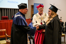 Kobieta w czarnej todze i birecie z logo uczelni podaje rękę dziekanowi w niebiesko-czarnej todze z łańcuchem. Obok stoi osoba w czerwonej todze z białym kołnierzem i birecie.