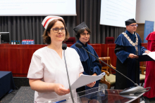 Uroczystość czepkowania absolwentów pielęgniarstwa i położnictwa. Kobieta w białym uniformie medycznym i czepku położnych stoi za mównicą, trzymając kartki papieru. Obok niej stoją dwie osoby w ciemnych togach akademickich.