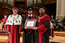 Trzech mężczyzn w togach akademickich stoi na scenie. Środkowy trzyma dyplom. Po lewej osoba w czerwonej todze z białym futrem i łańcuchem. Po prawej osoba w czerwono-czarnej todze z łańcuchem. W tle uczestnicy ceremonii w togach. Scena w eleganckim wnętrzu z drewnem i organami piszczałkowymi.