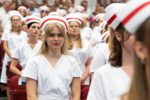Uroczystość czepkowania absolwentów pielęgniarstwa i położnictwa. Kobiety w białych uniformach medycznych i czepkach położnych stoją wzdłuż rzędów czerwonych krzeseł. Na pierwszym planie znajduje się kobieta z długimi blond włosami, patrząca przed siebie.